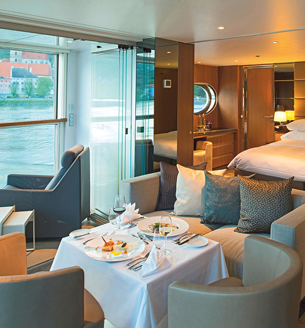  The Royal Panorama Suite showing a lounge area set up for in-suite dining, a queen bed and views of the passing river.