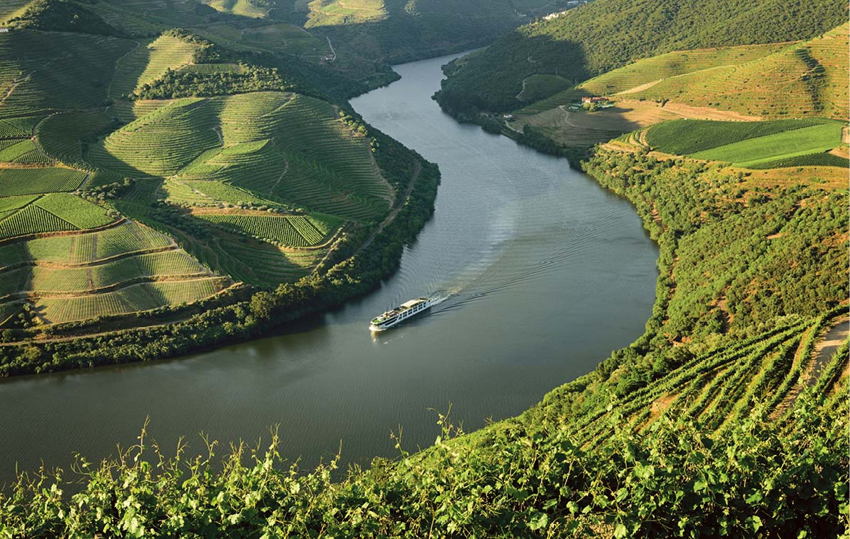 Bird’s eye shot of a white river cruise ship sailing through the Douro valley, with green fields and vineyards on either side 