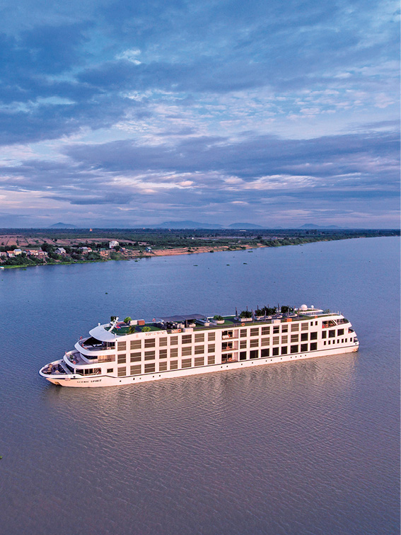 White cruise ship sailing on the Mekong river at sunset with cloudy blue skies and the shoreline with small houses in the distance 