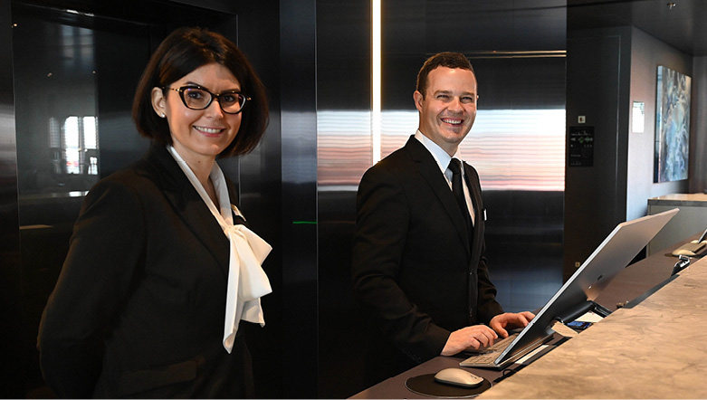 a male and a female member of reception staff on board Scenic Eclipse stand smiling at the desk ready to assist